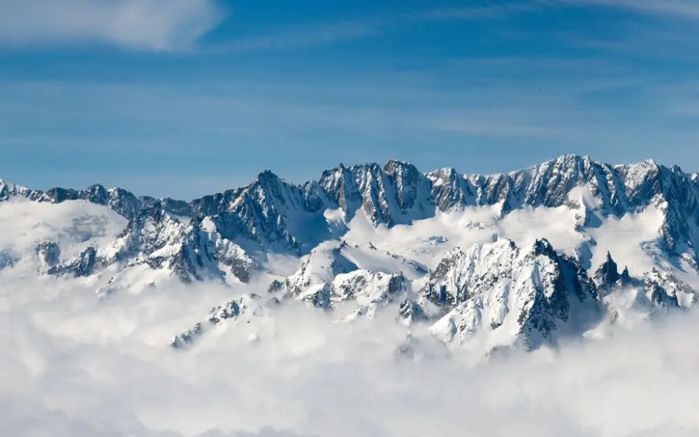 Symbolbild: Majestic snow-covered mountain peaks rise above clouds under a serene blue sky.
