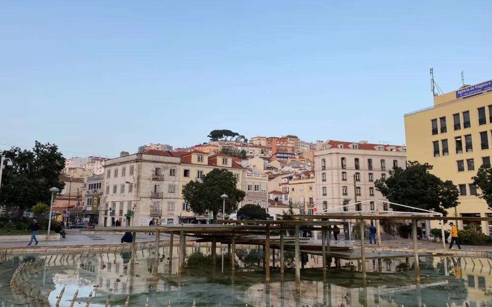 Symbolbild: A picturesque view of Lisbon's town square featuring buildings and a reflecting fountain under a clear sky.