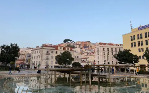 Symbolbild: A picturesque view of Lisbon's town square featuring buildings and a reflecting fountain under a clear sky.