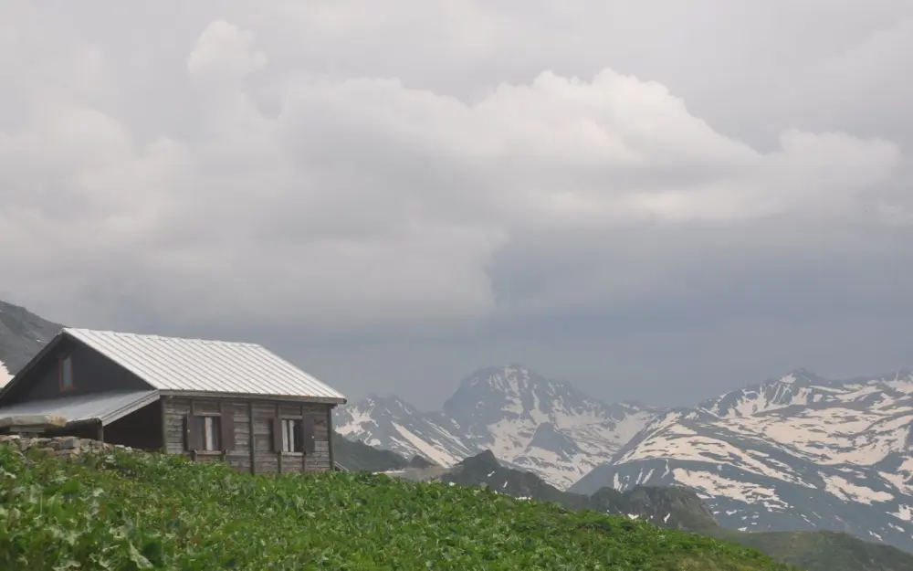 Symbolbild: A scenic view of a rustic wooden cabin surrounded by lush fields and snowcapped mountains.