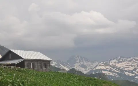 Symbolbild: A scenic view of a rustic wooden cabin surrounded by lush fields and snowcapped mountains.