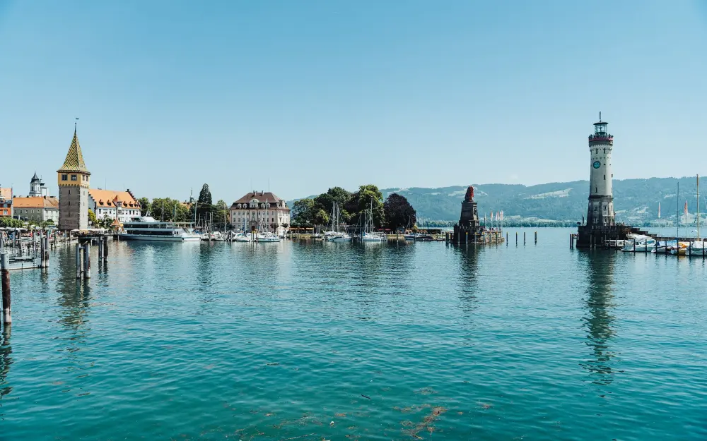 Panoramic view of Lindau harbor with lighthouse, boats, and buildings against blue sky.
