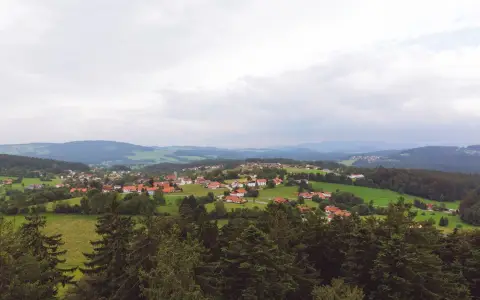 Symbolbild: Aerial view of a peaceful Bavarian village surrounded by lush greenery and rolling hills under a cloudy sky.