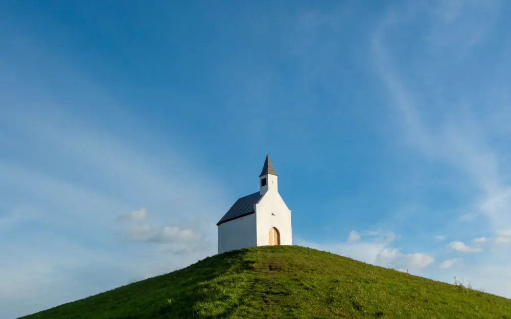 Symbolbild: A solitary white chapel with a steeple on a grassy hill under a clear blue sky.