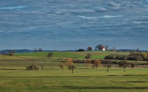 Symbolbild: Vast green fields and rolling hills with a small chapel under a clouded sky. Perfect rural tranquility.