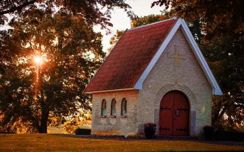 Symbolbild: A picturesque stone chapel with a red roof surrounded by trees at sunset, exuding tranquillity.