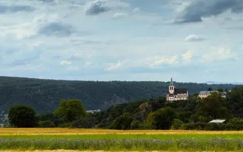 Symbolbild: Picturesque countryside scene with a church on a hill, surrounded by fields and trees under a cloudy sky.