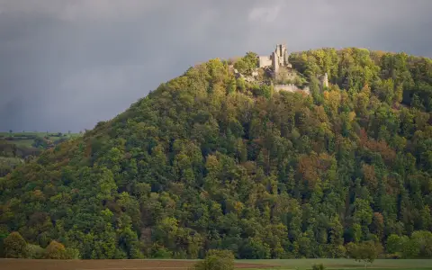 Symbolbild: Discover ancient castle ruins atop a lush, forested hill under dramatic skies.