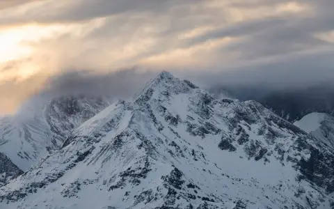 Symbolbild: A breathtaking view of a snow-covered mountain peak under a dawn sky with fog and clouds.