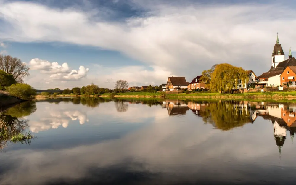 Symbolbild: Scenic view of a church and village reflecting on calm river waters in Höxter, Germany.
