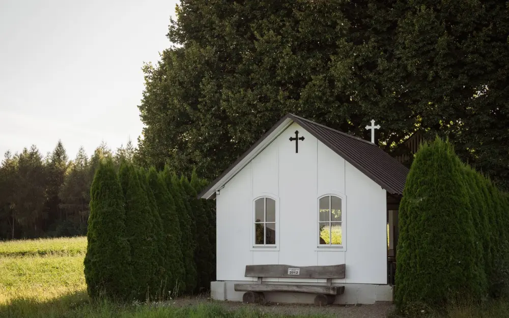Symbolbild: A peaceful small white church nestled among trees and greenery in a rural setting.