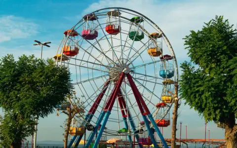 Symbolbild: Colorful Ferris wheel at an outdoor amusement park during daytime, surrounded by trees and blue sky.