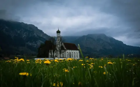Symbolbild: Scenic view of a historic church amidst a field of wildflowers with mountains in the background.