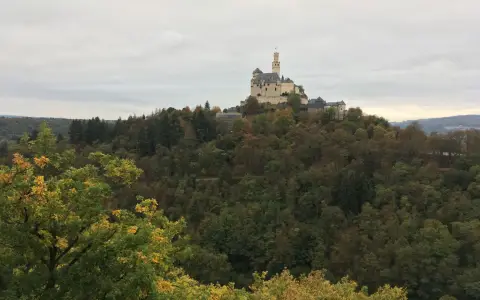 Symbolbild: Scenic view of Marksburg Castle atop a forested hill with vibrant fall colors under an overcast sky.