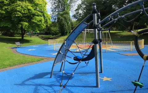 Symbolbild: A vibrant playground with blue rubber flooring and climbing structures in a lush green park.