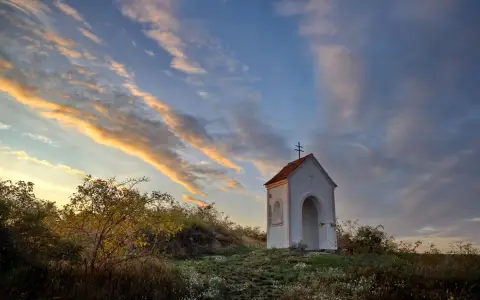 Symbolbild: A picturesque chapel stands on a hill under a vibrant evening sky, surrounded by nature.