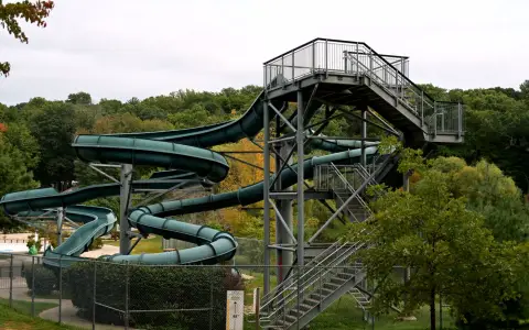 Symbolbild: A large, twisting outdoor water slide with a staircase, set in a forested amusement park, observed from a low angle.