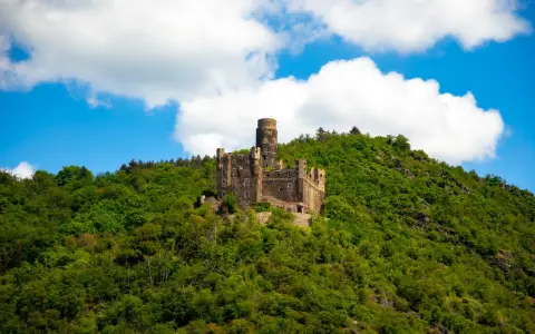 Symbolbild: Ancient castle atop a green hill surrounded by trees under a bright blue sky with clouds.