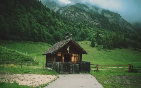 Symbolbild: A charming wooden cabin set in a lush green valley beneath misty mountains, showcasing rural serenity.