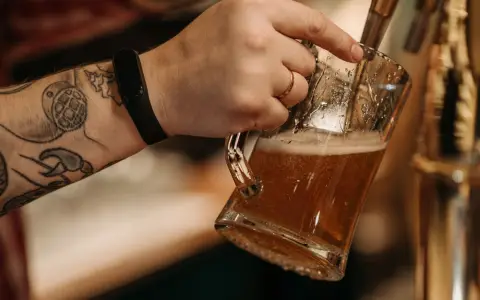Symbolbild: Close-up of a tattooed hand pouring beer into a glass mug from a draft tap.