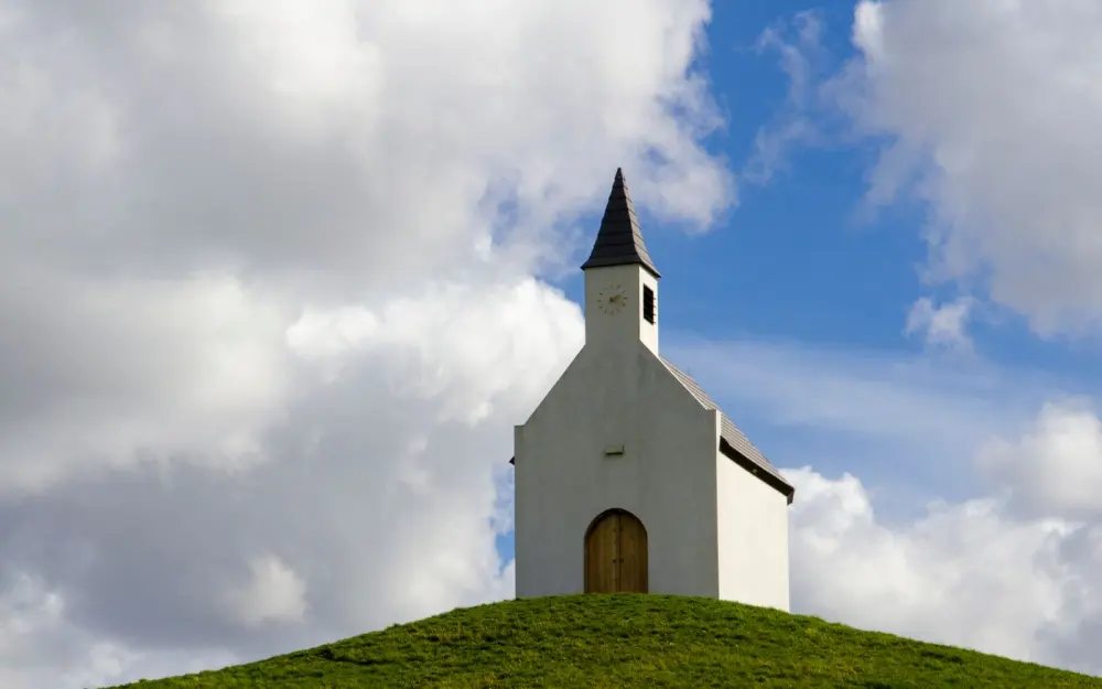 Symbolbild: A serene chapel perched on a grassy hilltop under a dramatic cloudy sky, capturing the essence of rural tranquility.