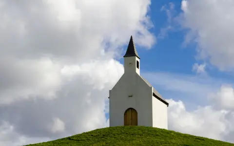 Symbolbild: A serene chapel perched on a grassy hilltop under a dramatic cloudy sky, capturing the essence of rural tranquility.